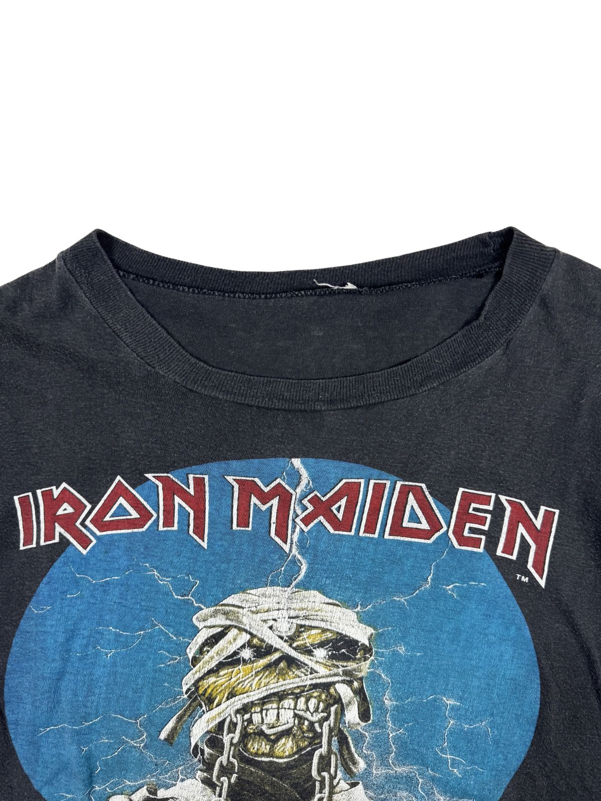 Nahaufnahme des Rundhalsausschnitts eines verwaschenen schwarzen Iron Maiden Shirts; im Anschnitt das ikonische Band-Logo und Eddie als Mumie.