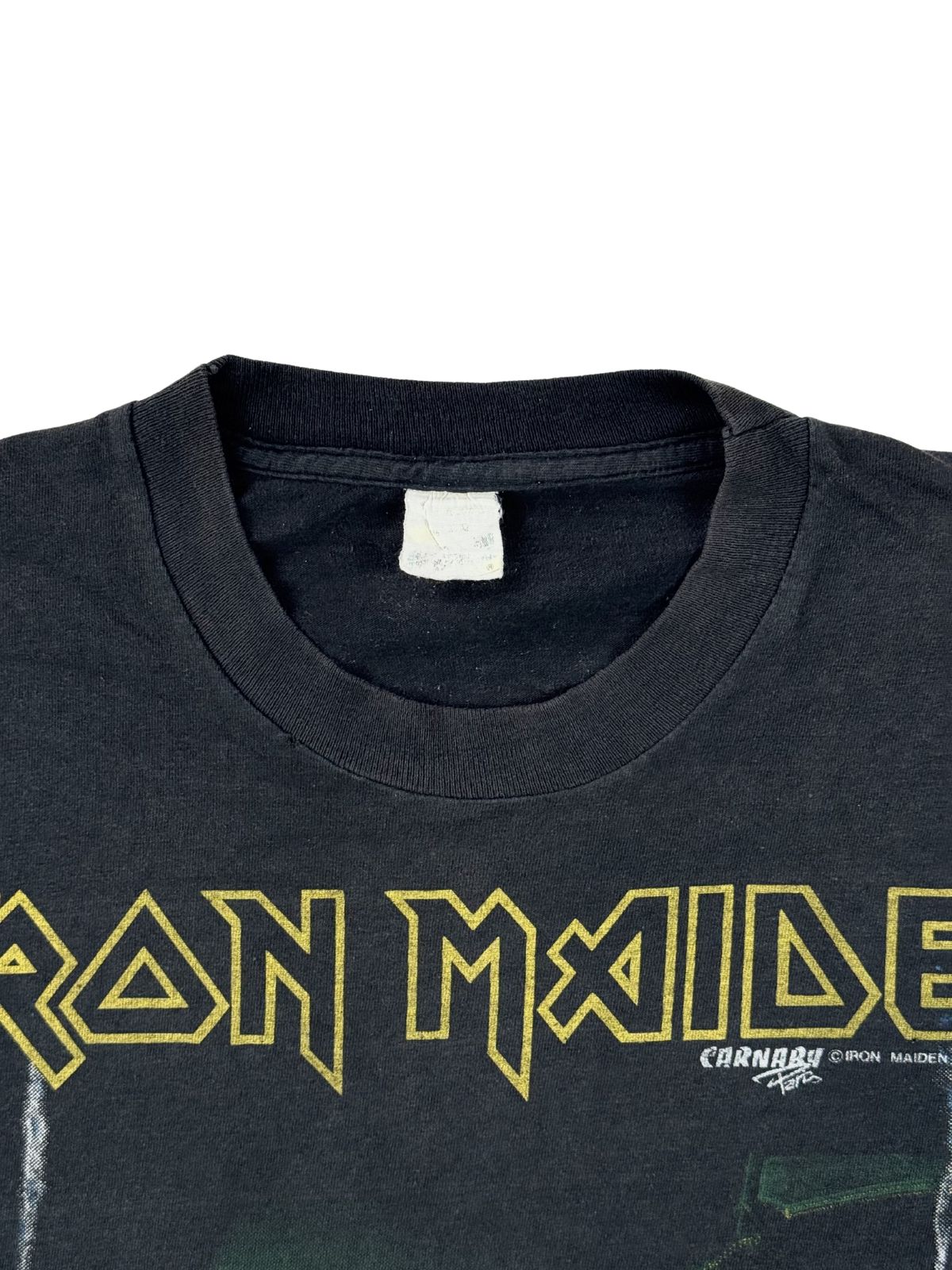 Detailansicht des Rundhalsausschnitts mit einem stark verwaschenen, weißen Nackenetikett direkt über dem gelben, klassischen Iron Maiden Logo-Schriftzug mit Carnaby-Lizenzierung.