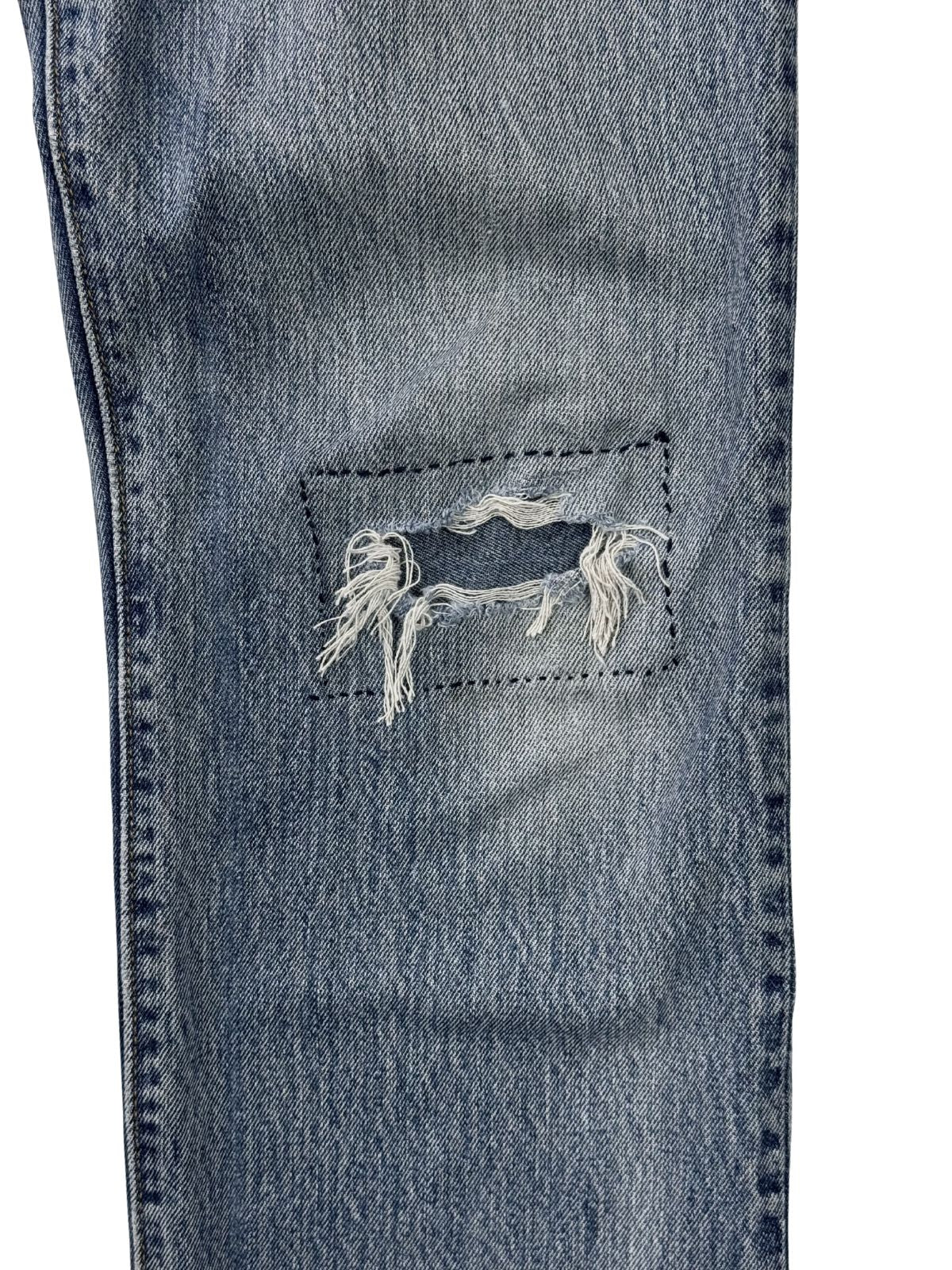Makroaufnahme einer handgenähten Reparaturstelle am Knie; zeigt dunkle Sticknähte um ein Loch im hellblauen Denim (Custom Repair Style).
