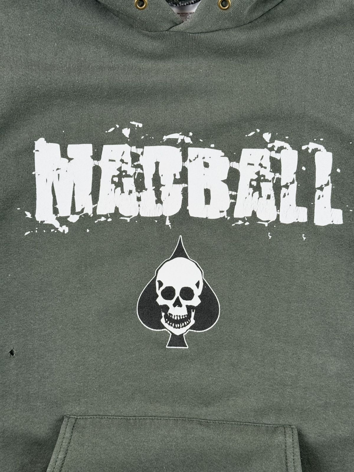 Nahaufnahme des vorderen Siebdrucks mit dem Madball-Schriftzug und dem Pik-As-Totenkopf. Links unterhalb des Prints ist ein kleines Loch (Pinhole) im grünen Sweatstoff erkennbar.