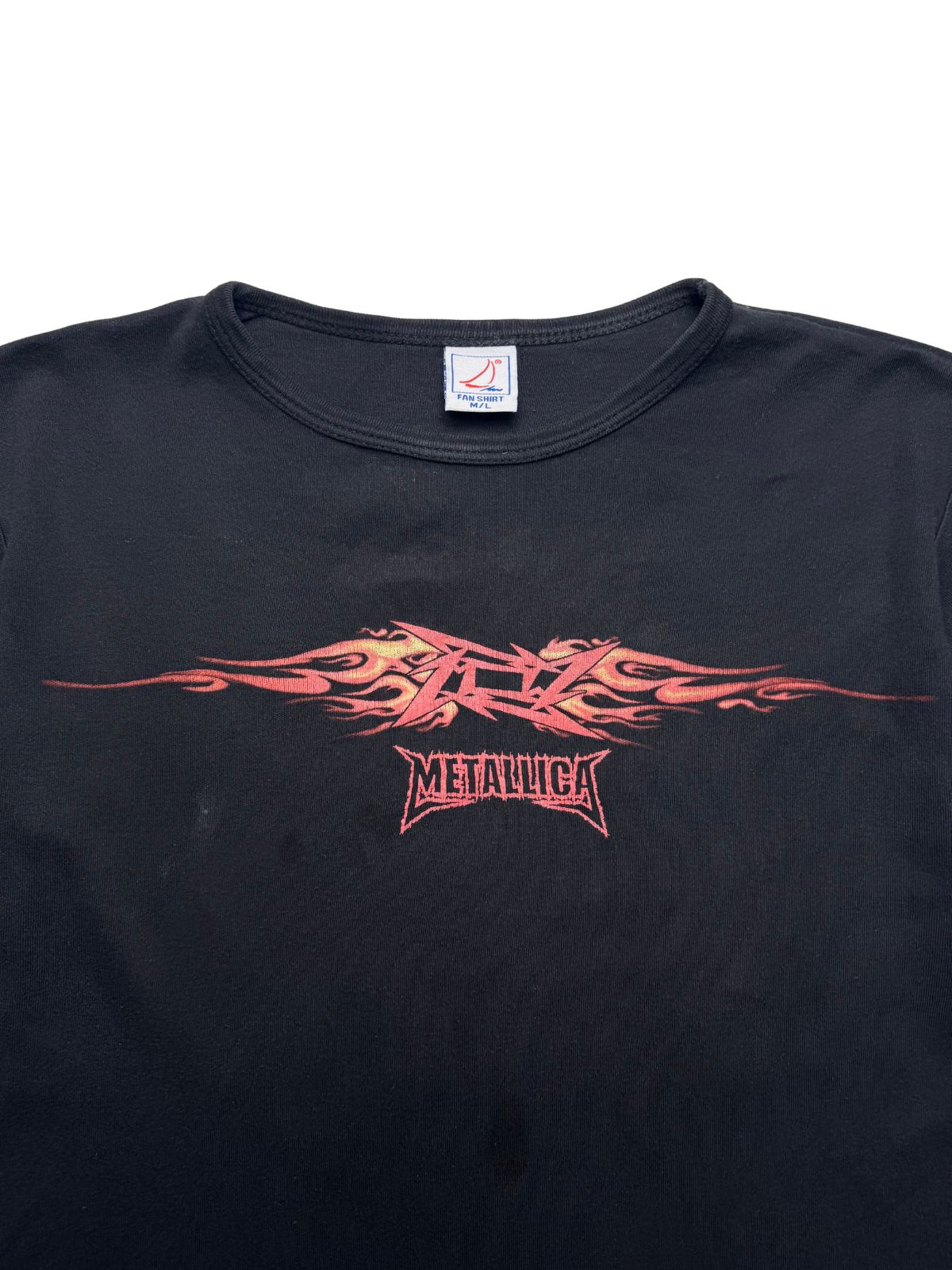 Detailansicht der vorderen Brustpartie mit weitem Rundhalsausschnitt, dem „FAN SHIRT“-Nackenetikett und dem rot-gelben Metallica-Flammen-Grafikdruck.