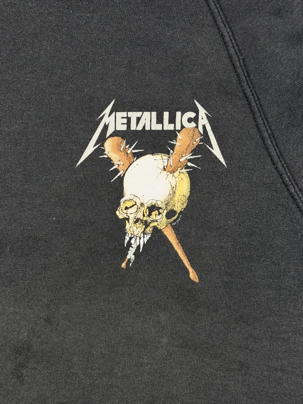 Detailansicht des Brust-Grafikdrucks: Ein gelblich-weißer Totenkopf mit zwei gekreuzten, dornigen Knochen hinter dem Metallica-Logo.