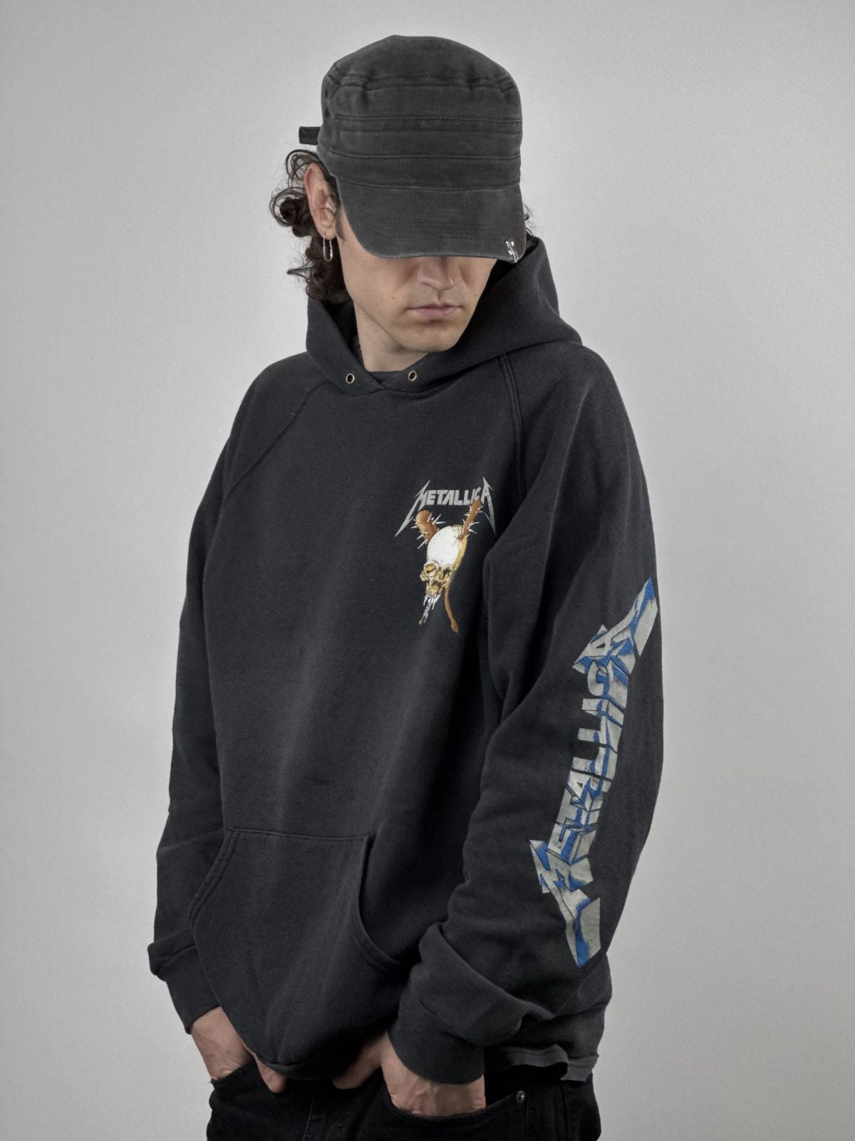 Ein männliches Model mit lockigem Haar und verwaschener schwarzer Schirmmütze trägt den Vintage Metallica Hoodie; zu sehen sind der Brust-Totenkopf-Print, die Kängurutasche und das Logo am Ärmel.