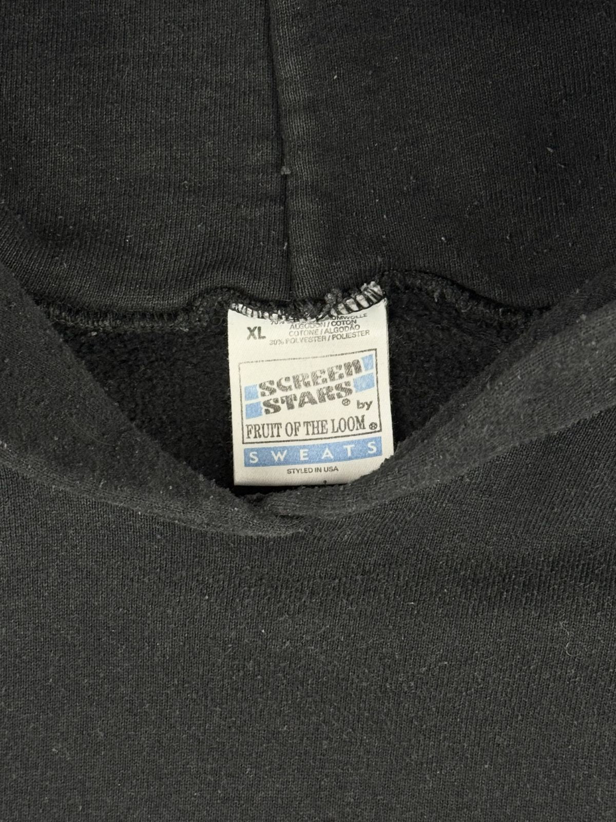 Makroaufnahme des Nackenetiketts des schwarzen 94er Metallica-Hoodies. Das weiße Stofflabel zeigt das blau-graue „Screen Stars by Fruit of the Loom“-Logo mit dem Zusatz „SWEATS – Styled in USA“ sowie die Angabe der Größe XL und Materialhinweise.