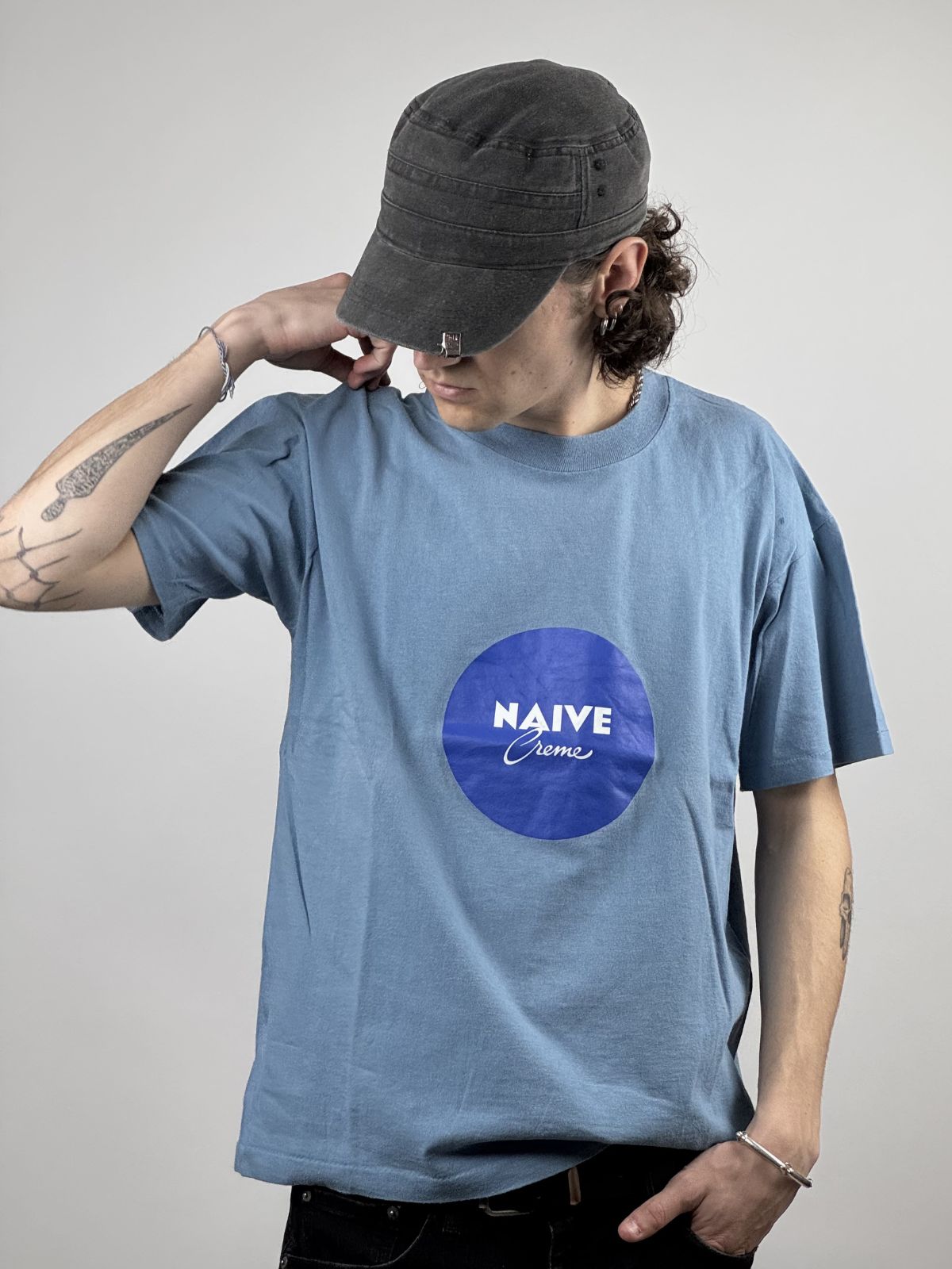 Ein Model trägt das hellblaue Naive-Parodie-Shirt in Kombination mit einer grauen Mütze; Fokus auf dem Grafik-Brustprint und der lockeren Passform.