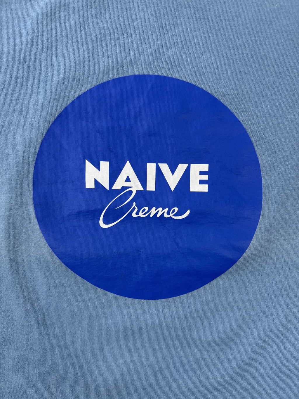 Hochauflösende Detailansicht des Brustprints: Dunkelblauer Kreis mit weißem „NAIVE Creme“ Schriftzug, der das Nivea-Design parodiert.