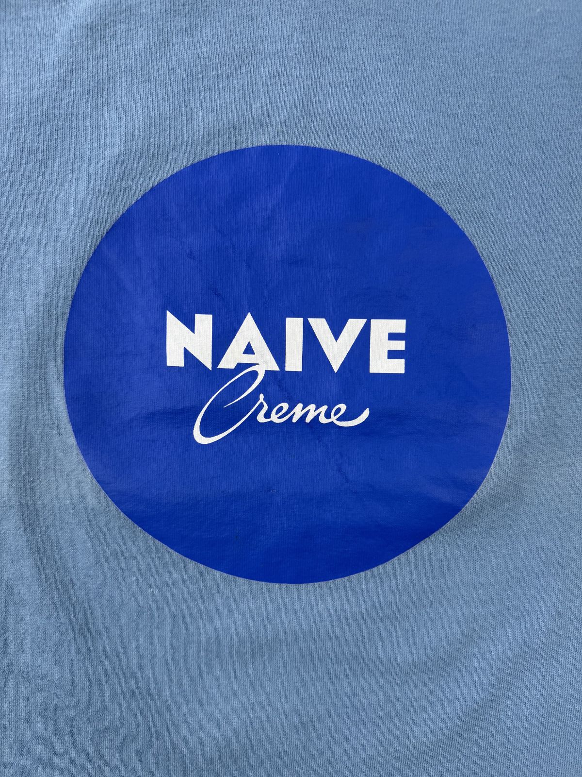 Hochauflösende Detailansicht des Brustprints: Dunkelblauer Kreis mit weißem „NAIVE Creme“ Schriftzug, der das Nivea-Design parodiert.