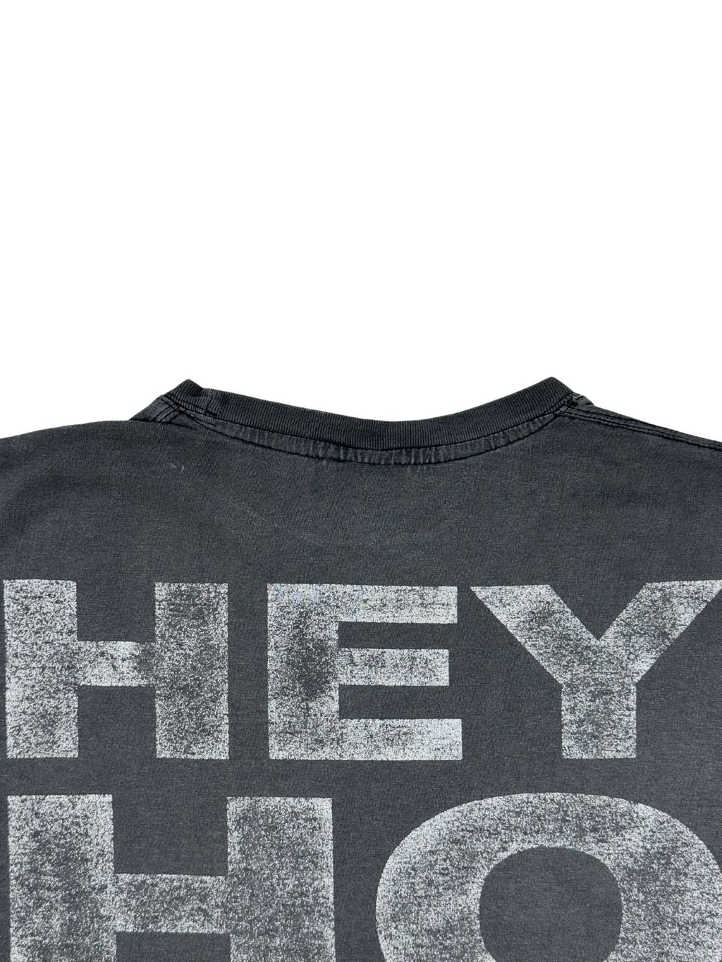 Nahezu-Ausschnitt der Rückseite des Shirts, der die rissige, verwaschene Textur des weißen „HEY HO“-Aufdrucks auf dem dunkelgrauen Baumwollstoff zeigt.
