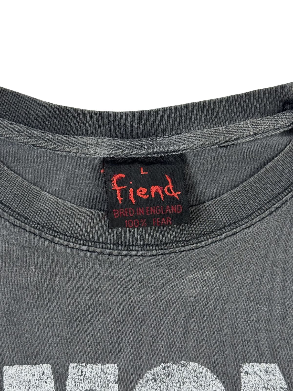 Makroaufnahme des Nackenetiketts eines 90er-Jahre Vintage-Shirts. Das schwarze Stofflabel zeigt in roter Schrift die Marke „Fiend“, die Größe L sowie den Text „Bred in England – 100% Fear“.