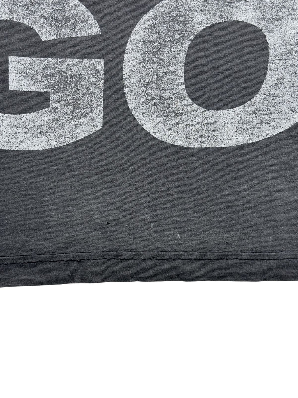 Nahaufnahme einer offenen bzw. beschädigten Nahtstelle am unteren Saum des grauen Vintage-T-Shirts, direkt neben einem kleinen Loch im Stoff.