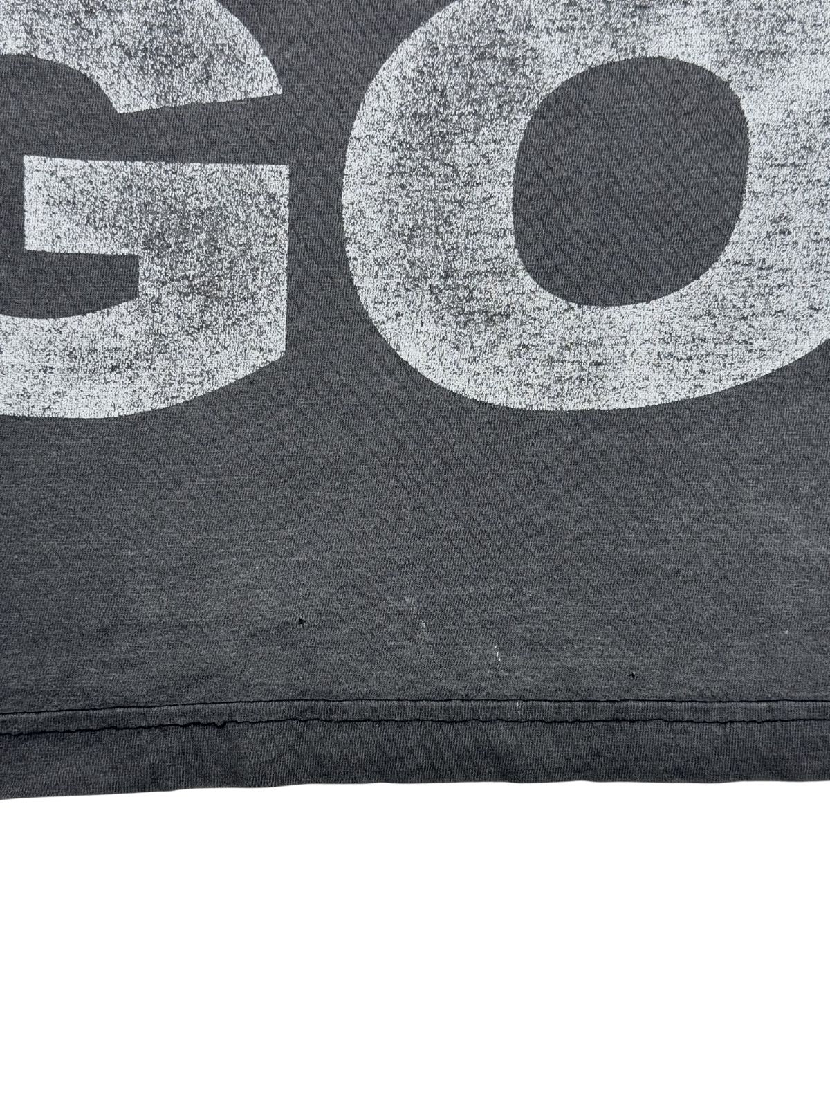Nahaufnahme einer offenen bzw. beschädigten Nahtstelle am unteren Saum des grauen Vintage-T-Shirts, direkt neben einem kleinen Loch im Stoff.