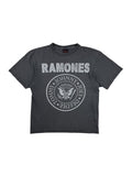Nahaufnahme der Vorderseite eines grauen Vintage Ramones T-Shirts aus den 1990er Jahren mit dem klassischen runden Band-Logo (Adler mit Baseballschläger und den Namen Johnny, Joey, Dee Dee, Tommy) und weißem Ramones-Schriftzug. Flatlay auf weißem Hintergrund.