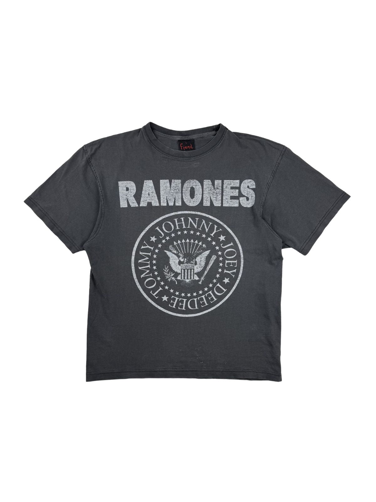 Nahaufnahme der Vorderseite eines grauen Vintage Ramones T-Shirts aus den 1990er Jahren mit dem klassischen runden Band-Logo (Adler mit Baseballschläger und den Namen Johnny, Joey, Dee Dee, Tommy) und weißem Ramones-Schriftzug. Flatlay auf weißem Hintergrund.