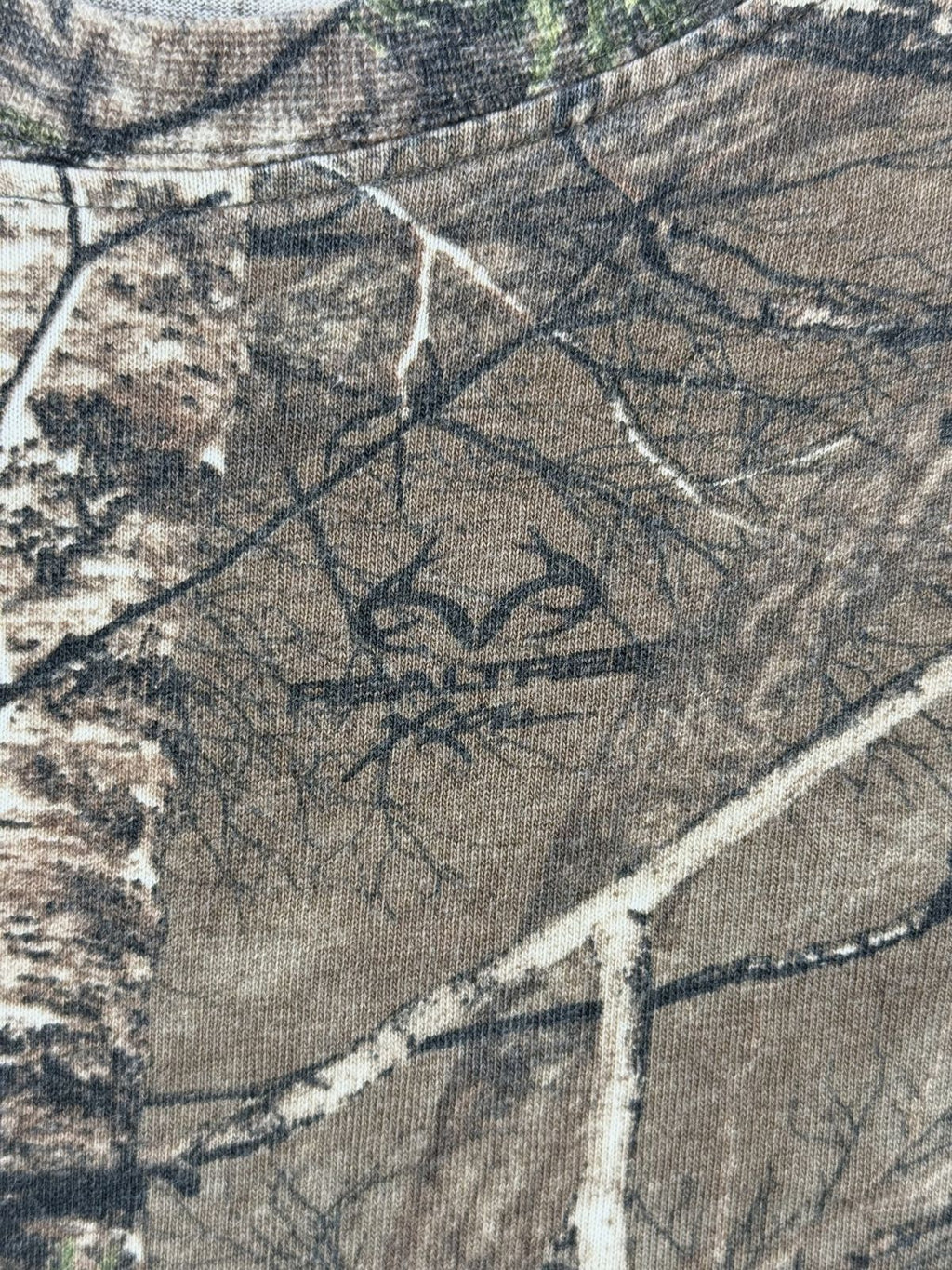 Makroaufnahme des im Stoff eingearbeiteten, originalen „Realtree Xtra“ Markenlogos inmitten des waldigen Camouflage-Musters.