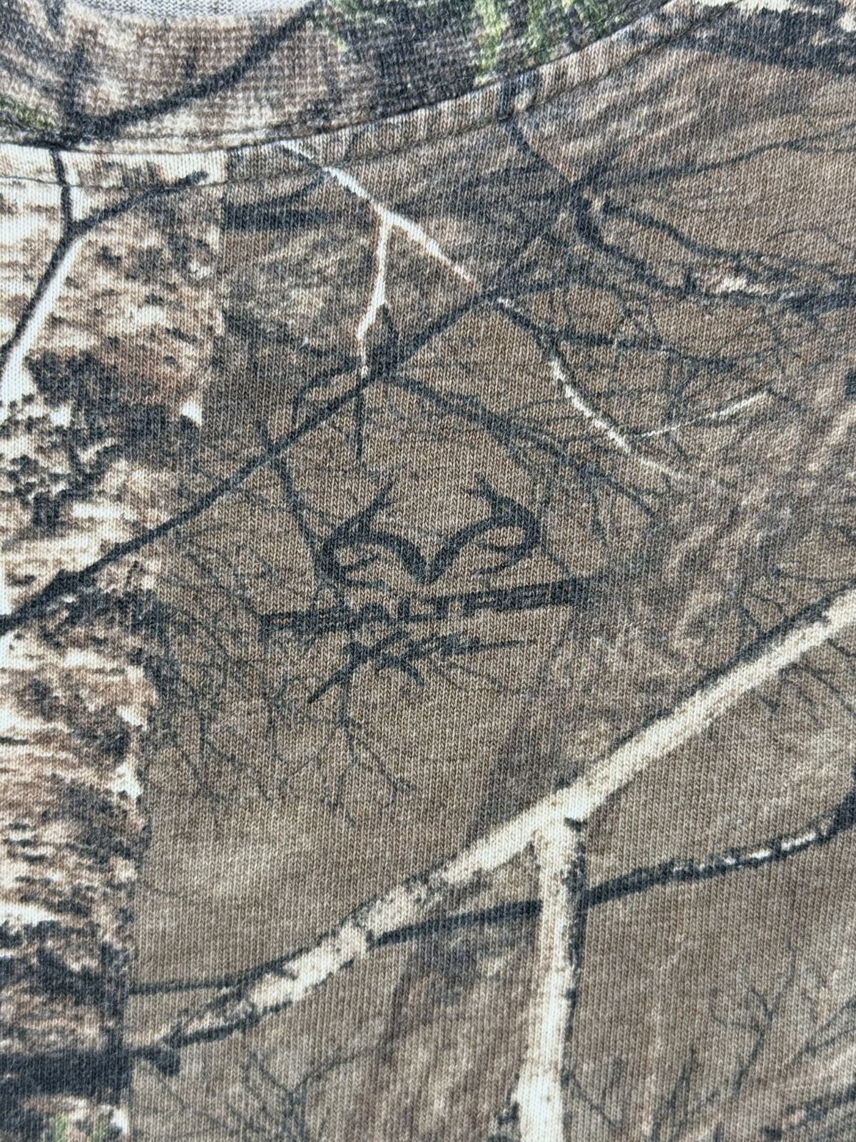 Makroaufnahme des im Stoff eingearbeiteten, originalen „Realtree Xtra“ Markenlogos inmitten des waldigen Camouflage-Musters.