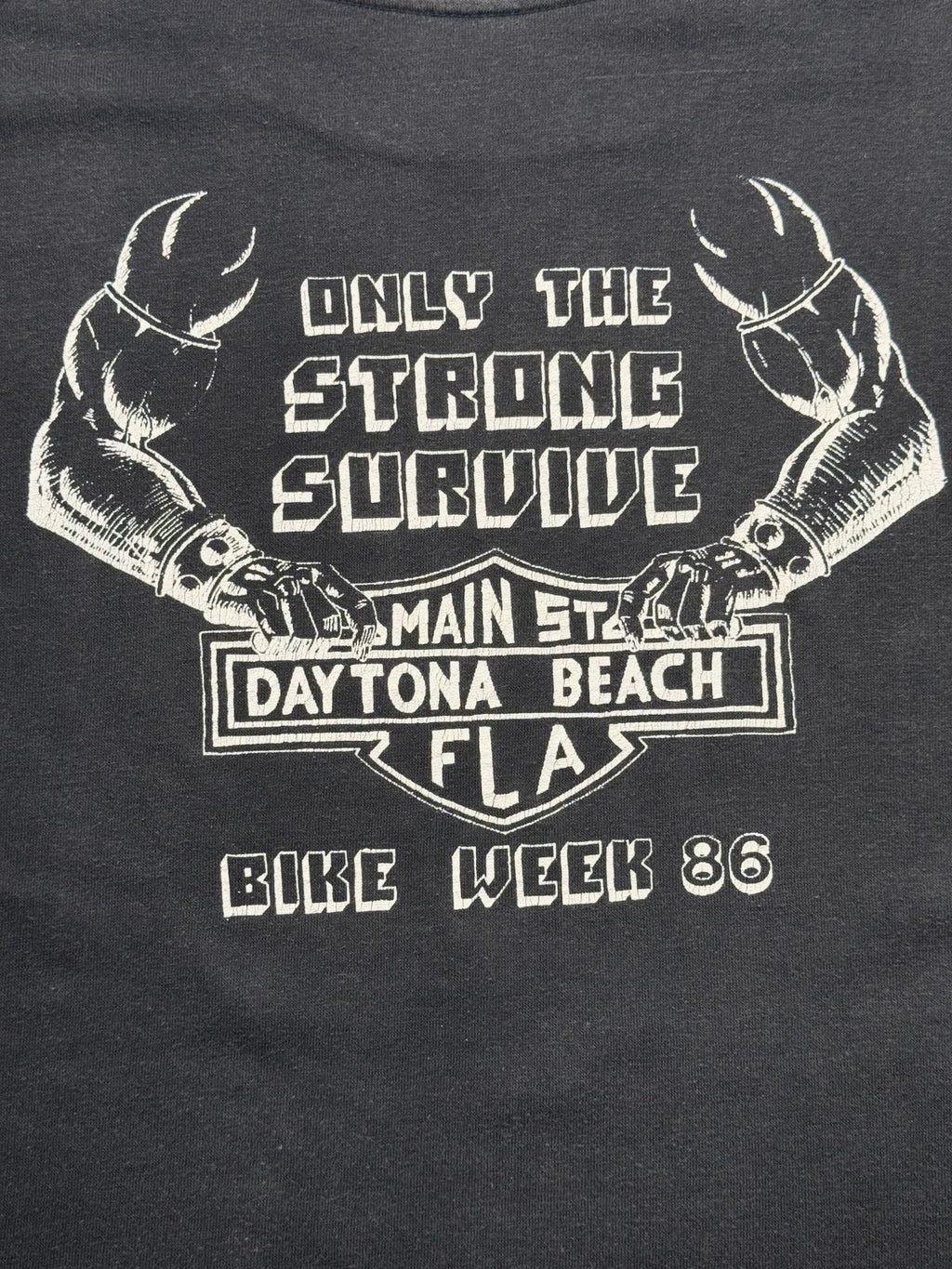 Detaillierte Ansicht des Rückenprints mit muskulösen Biker-Armen, Nietenbändern, Harley-Schild-Emblem und dem Slogan „ONLY THE STRONG SURVIVE - BIKE WEEK 86“.