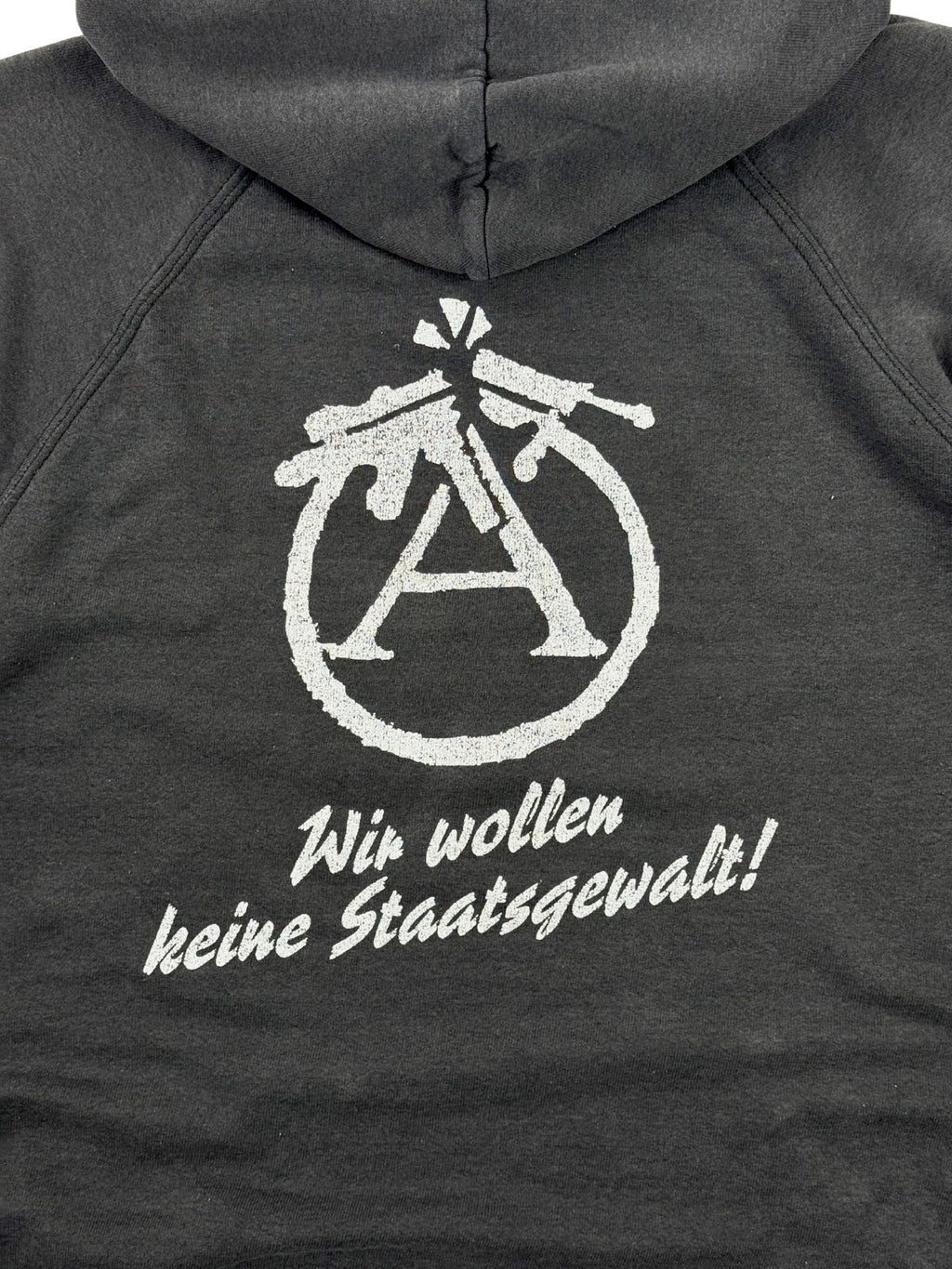 Nahaufnahme des politischen Rückendrucks. Zu sehen ist ein abgewandeltes Anarchie-A-Zeichen, bei dem der Kreis aus zwei zerbrochenen Gewehren gebildet wird, darunter der weiße Schriftzug „Wir wollen keine Staatsgewalt!“.