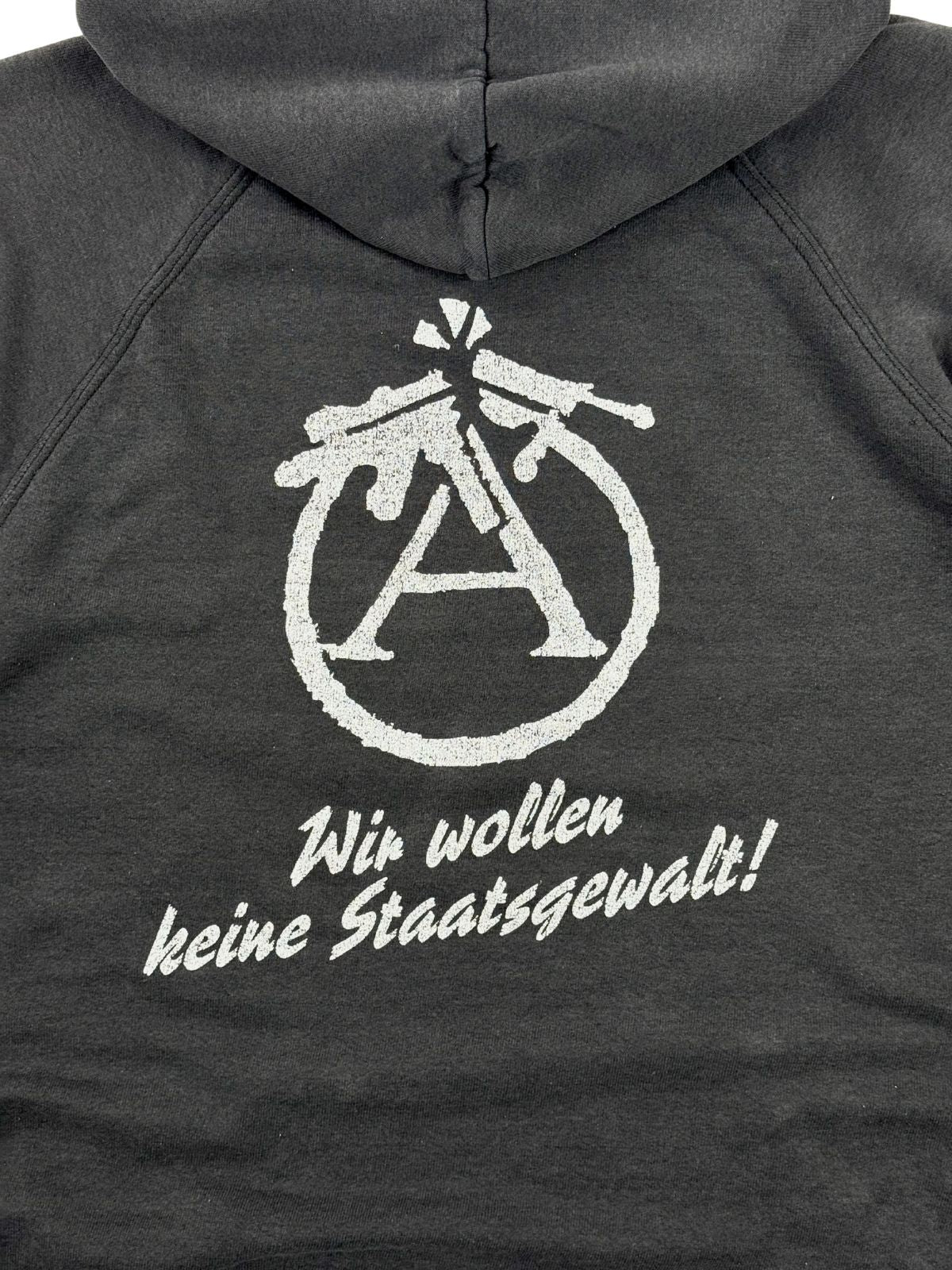 Nahaufnahme des politischen Rückendrucks. Zu sehen ist ein abgewandeltes Anarchie-A-Zeichen, bei dem der Kreis aus zwei zerbrochenen Gewehren gebildet wird, darunter der weiße Schriftzug „Wir wollen keine Staatsgewalt!“.