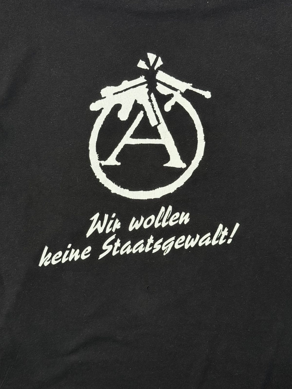 Nahaufnahme des weißen Backprints mit der politischen Parole „Wir wollen keine Staatsgewalt!“ unter dem modifizierten, antimilitaristischen Anarchie-A-Symbol.