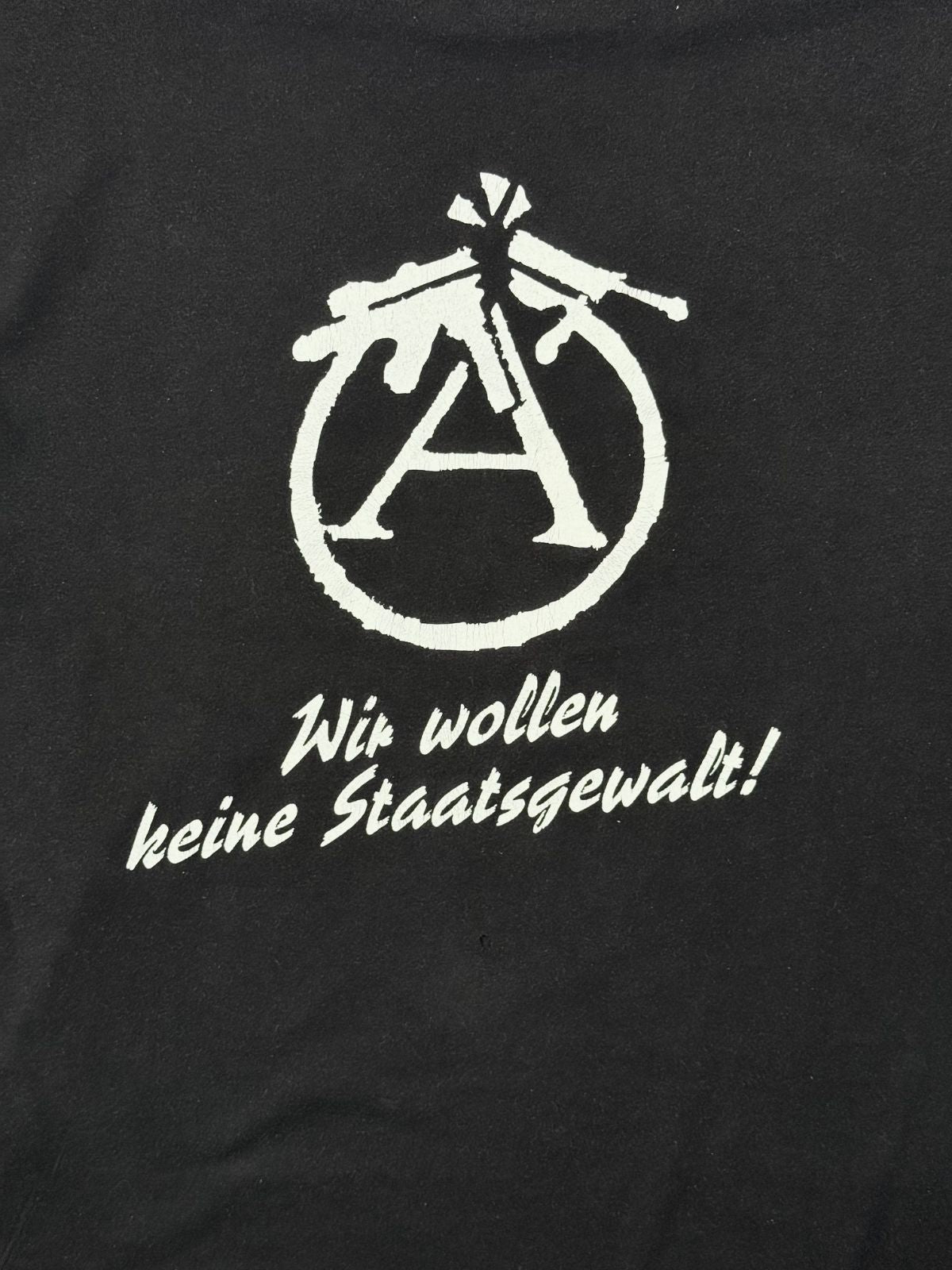 Nahaufnahme des weißen Backprints mit der politischen Parole „Wir wollen keine Staatsgewalt!“ unter dem modifizierten, antimilitaristischen Anarchie-A-Symbol.