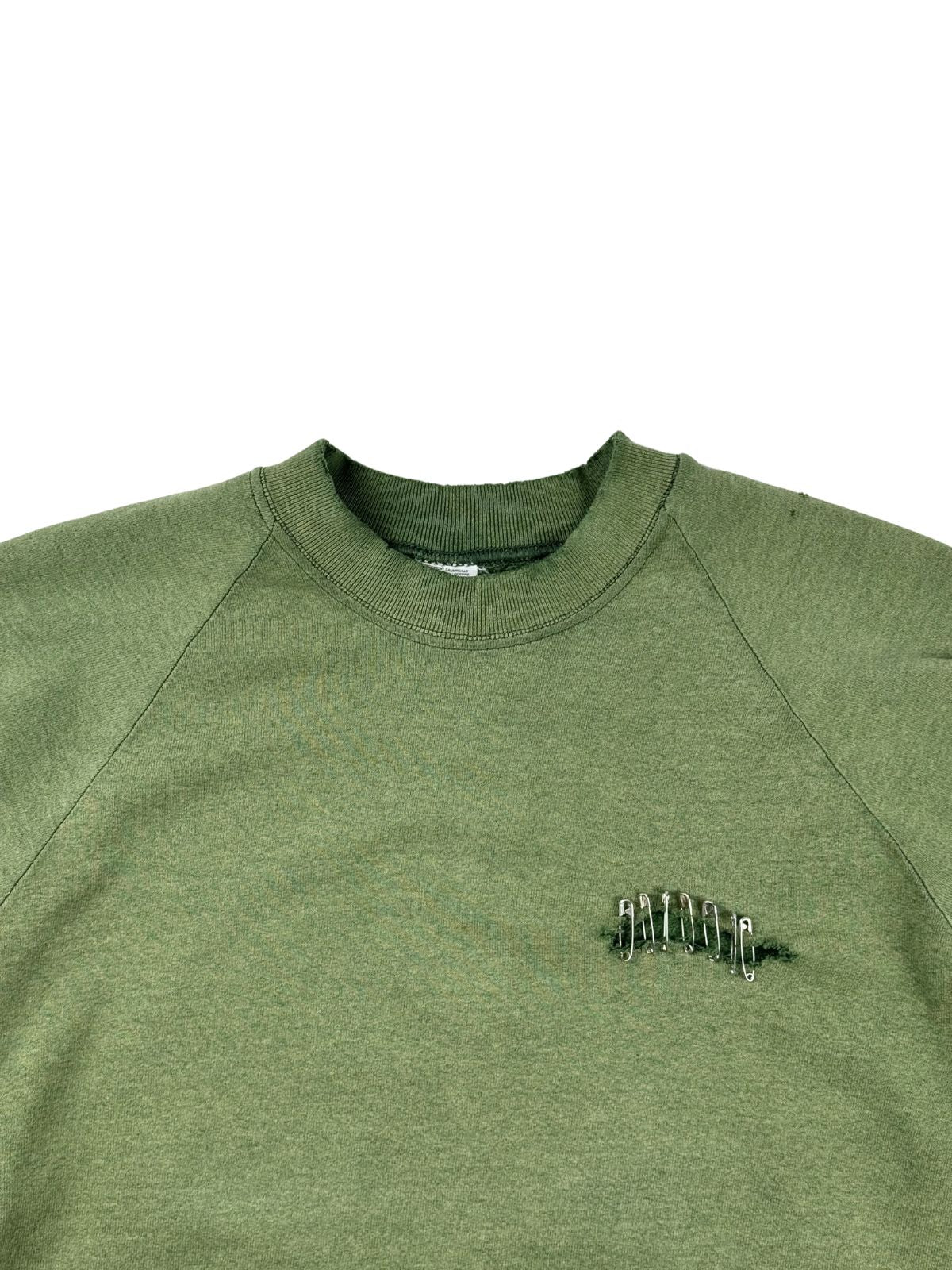 Detailansicht der Vorderseite des olivgrünen Pullovers mit Fokus auf den gerippten Rundhalsausschnitt und den mit Sicherheitsnadeln fixierten Riss.