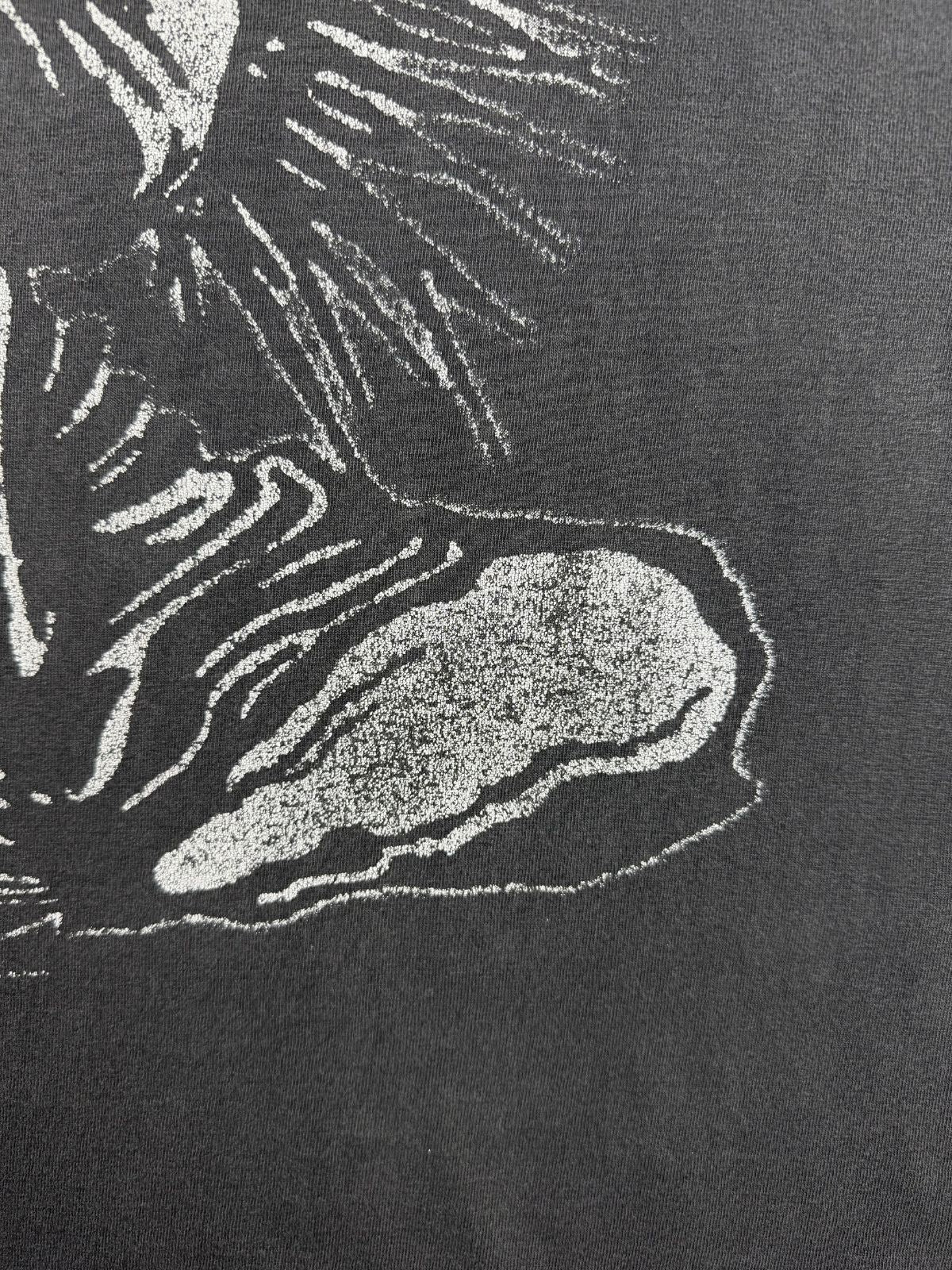 Detailansicht des vorderen Siebdrucks auf dem verwaschenen Baumwollstoff, die den unteren Teil der zerschlissenen Totenkopf-Grafik im distressed Look zeigt.