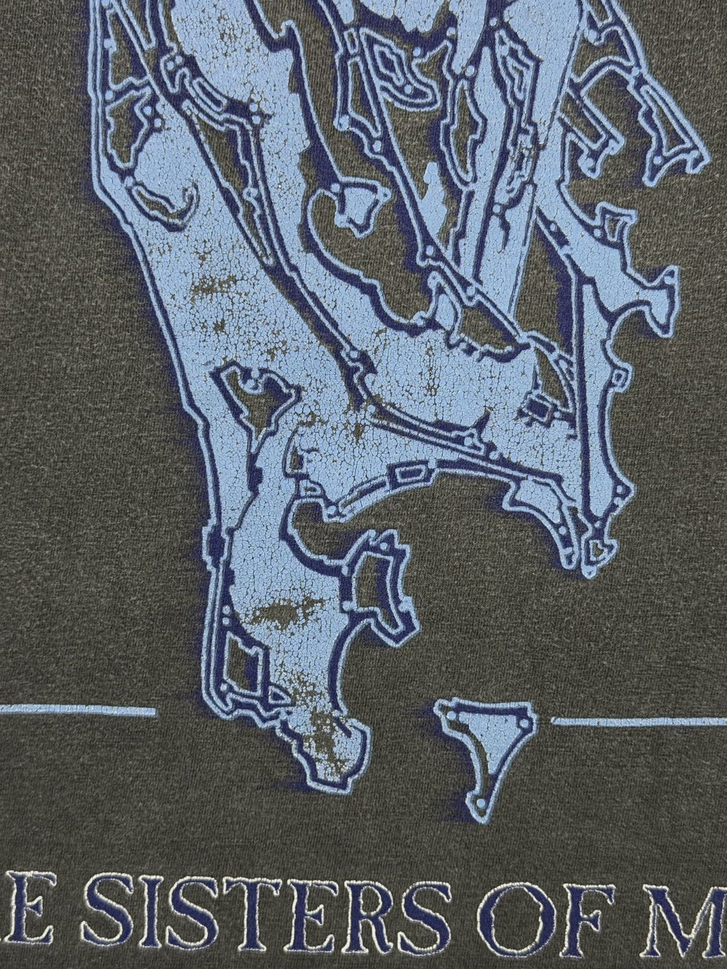 Detaillierte Makroaufnahme des verwitterten Frontprints auf einem Vintage-Sisters-of-Mercy-T-Shirt. Zu sehen ist die ausgeprägte Rissbildung (Cracking) im hellblauen Siebdruck des abstrakten Motivs auf dem dunkelgrauen Baumwollstoff. Unten im Bild sind die Reste der Schrift „THE SISTERS OF M“ zu erkennen. Dieses Foto verdeutlicht den authentischen Alterungsprozess und den Used-Look des originalen Merchandise-Artikels von 1993.