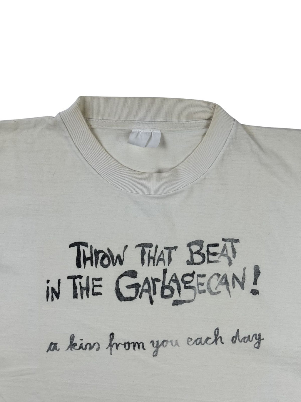 Detailansicht des Ausschnitts mit dem schwarzen Print „THROW THAT BEAT IN THE GARBAGECAN! a kiss from you each day“ in verspielter Handschrift-Optik. Ein verwaschenes, weißes Stoffetikett ist am Kragen erkennbar.