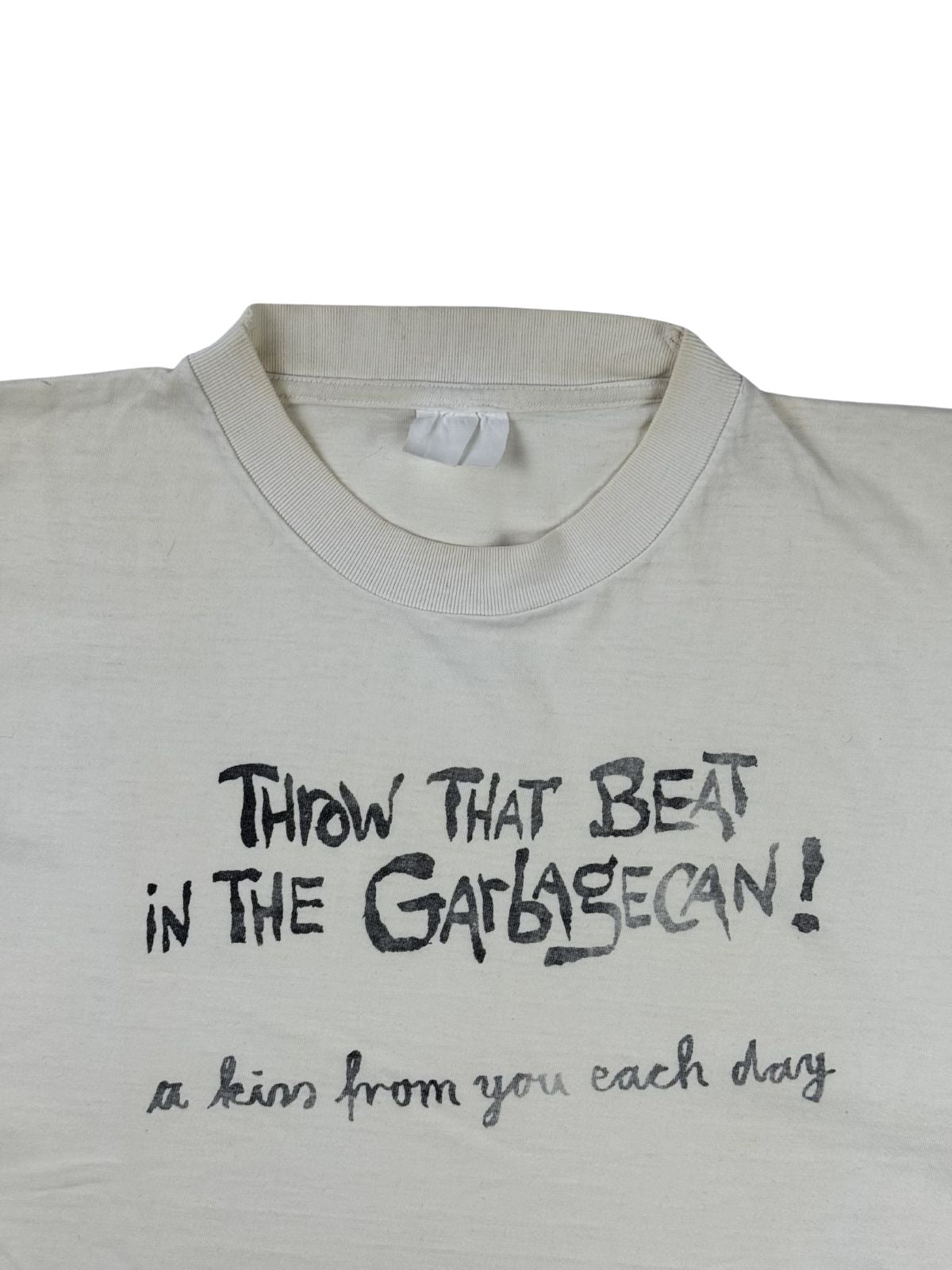 Detailansicht des Ausschnitts mit dem schwarzen Print „THROW THAT BEAT IN THE GARBAGECAN! a kiss from you each day“ in verspielter Handschrift-Optik. Ein verwaschenes, weißes Stoffetikett ist am Kragen erkennbar.