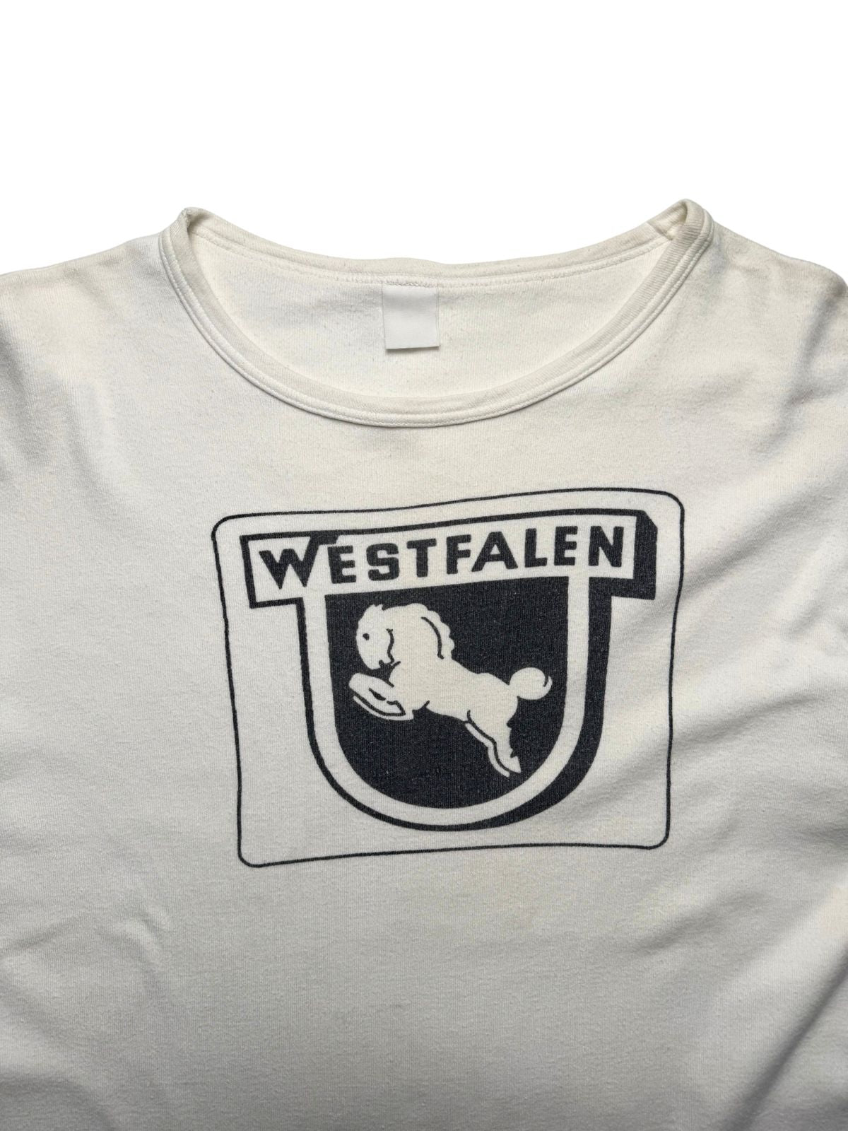 Detailansicht der vorderen Brustpartie mit weitem Rundhalsausschnitt, blanko Nackenetikett und dem schwarzen Siebdruck-Wappen mit Schriftzug „WESTFALEN“.